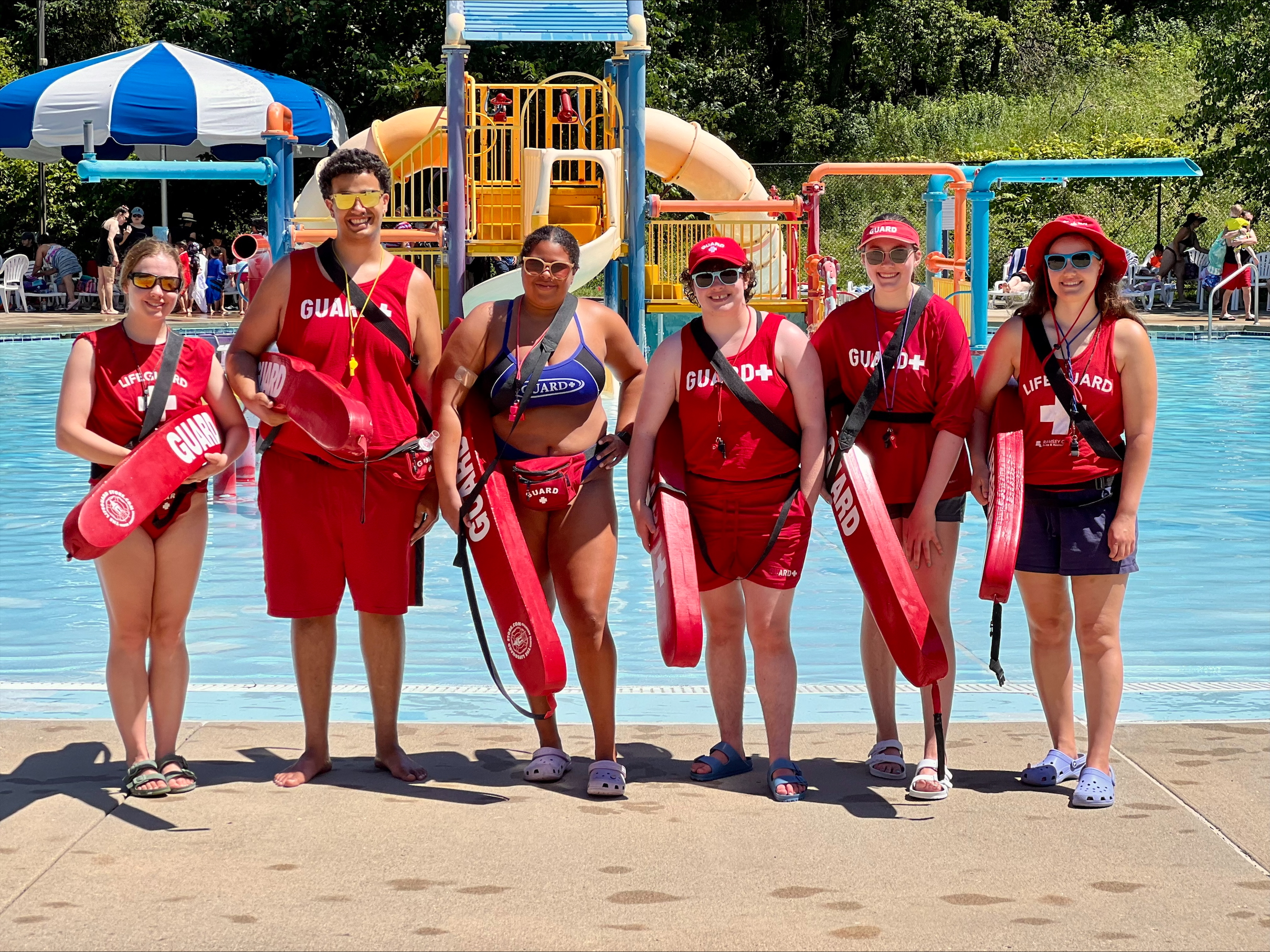 Six smiling lifeguards in red and sunglasses standing in front of a pool.