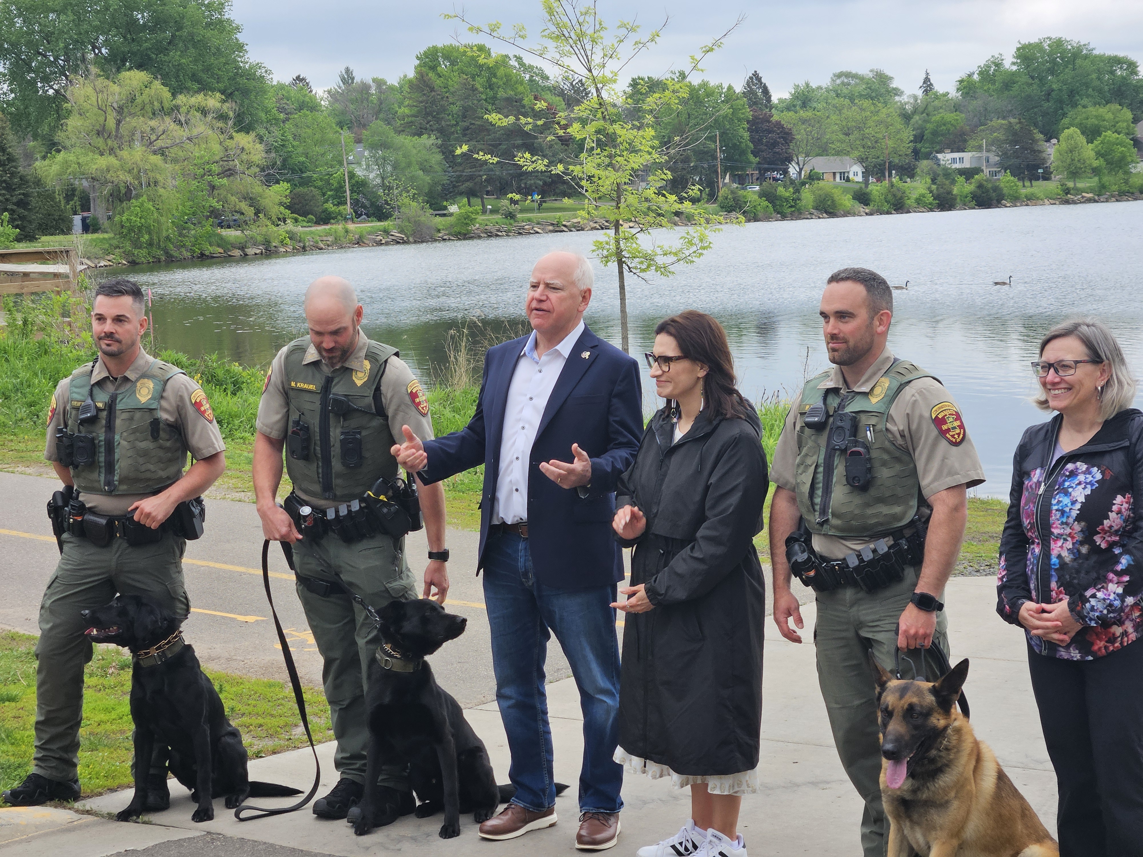 Govenor Walz, Lieutenant Governor Flanagan, DNR Commissioner Sarah Strommen, and four DNR officers along with their DNR sniffing dogs are posed in a photo in front of a lake. 