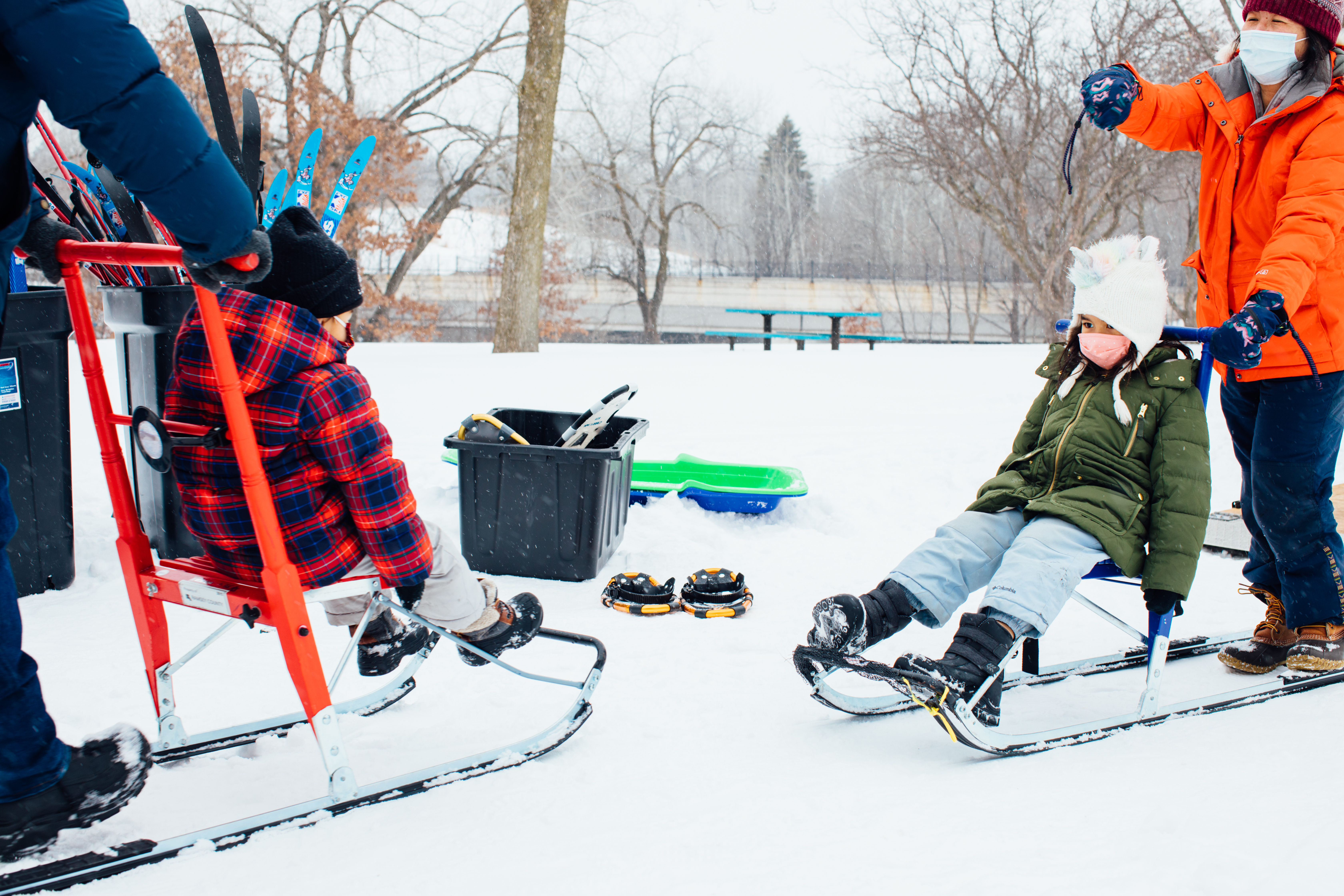 During winter, two adults pushing children on kick-sleds in the snow.