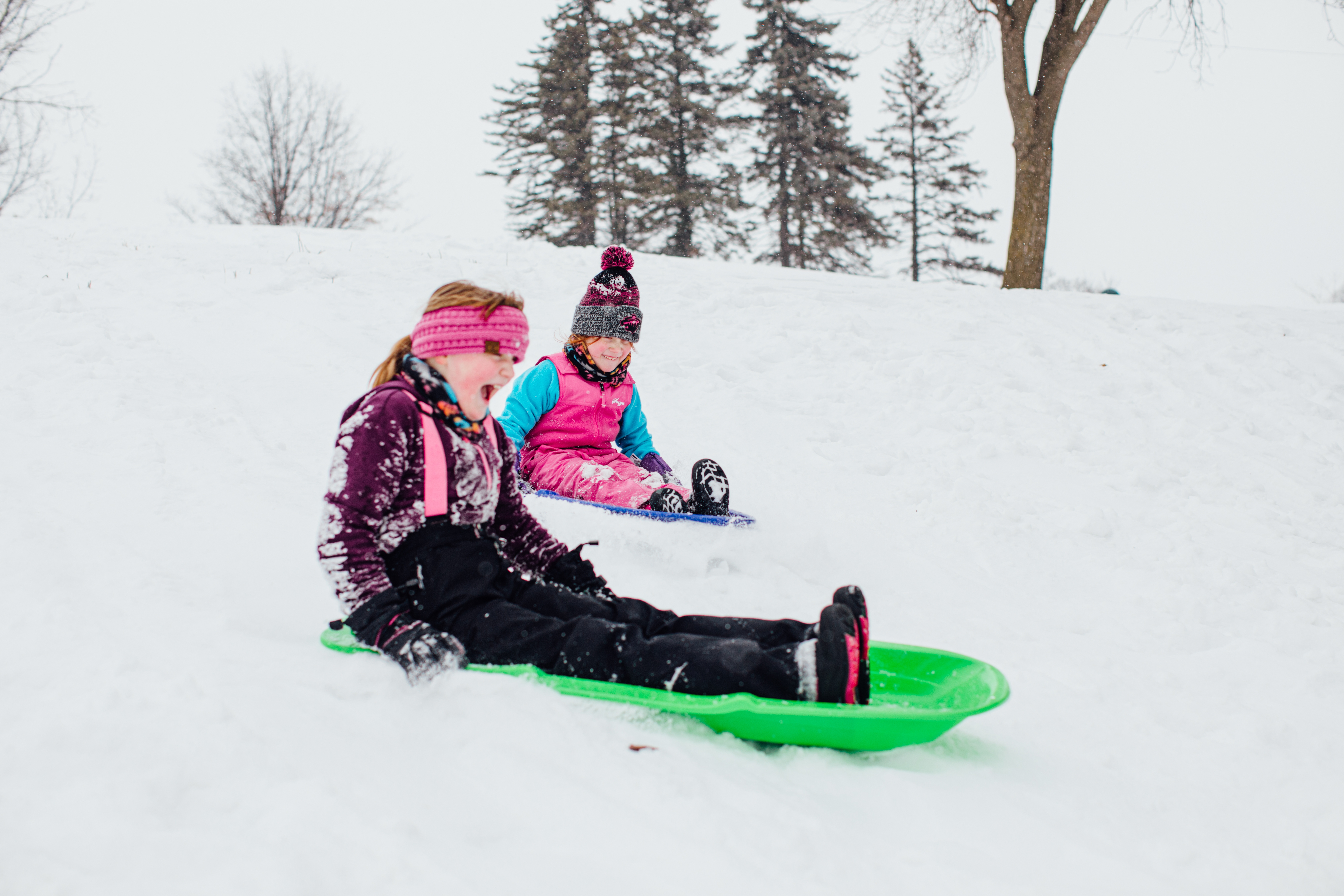 In the winter, two girls sitting on their sleds on the snow hill.