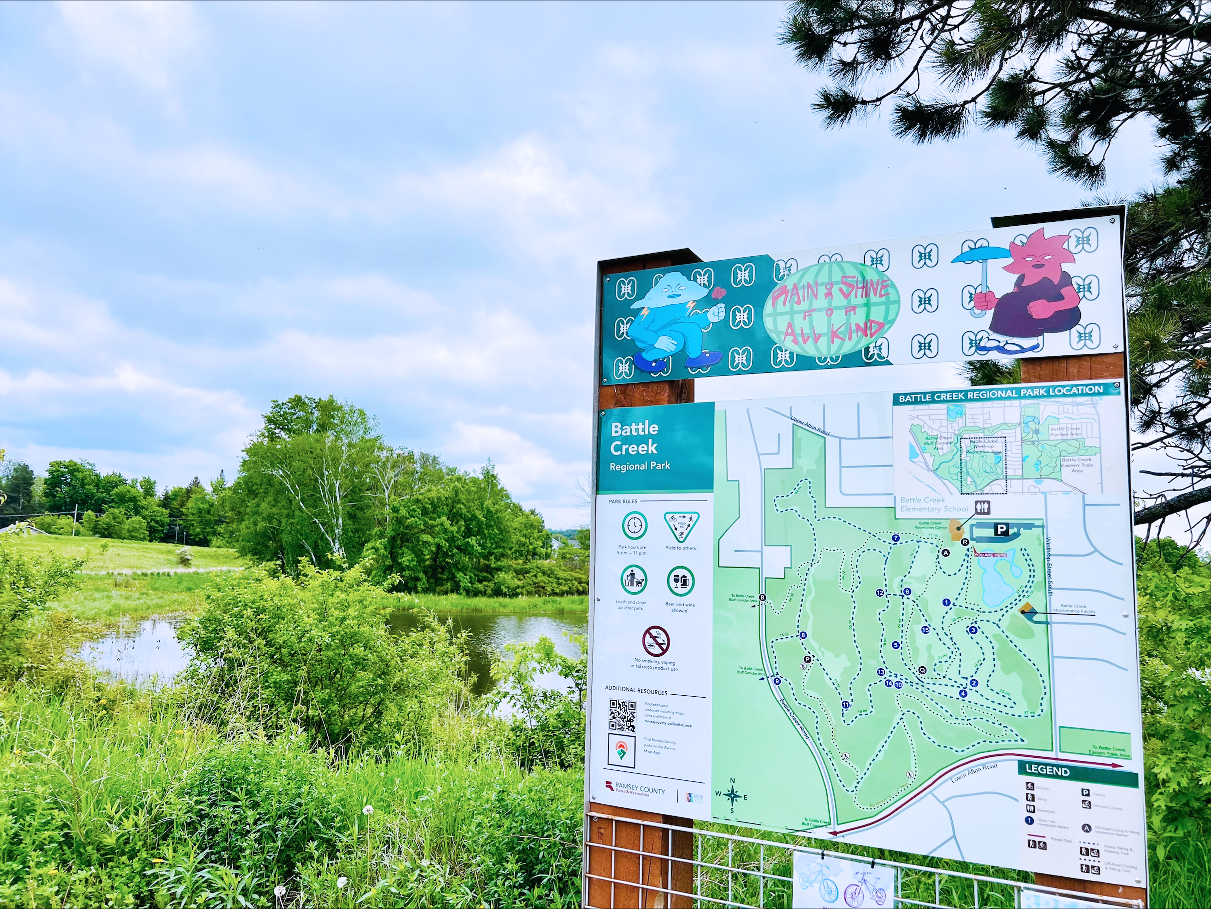 Battle Creek Recreation Trail sign that depicts a map of the area. On top of the map is artwork done by CIRCE of a red and blue caricature on either side with the words in the middle saying "rain or shine for all kind."