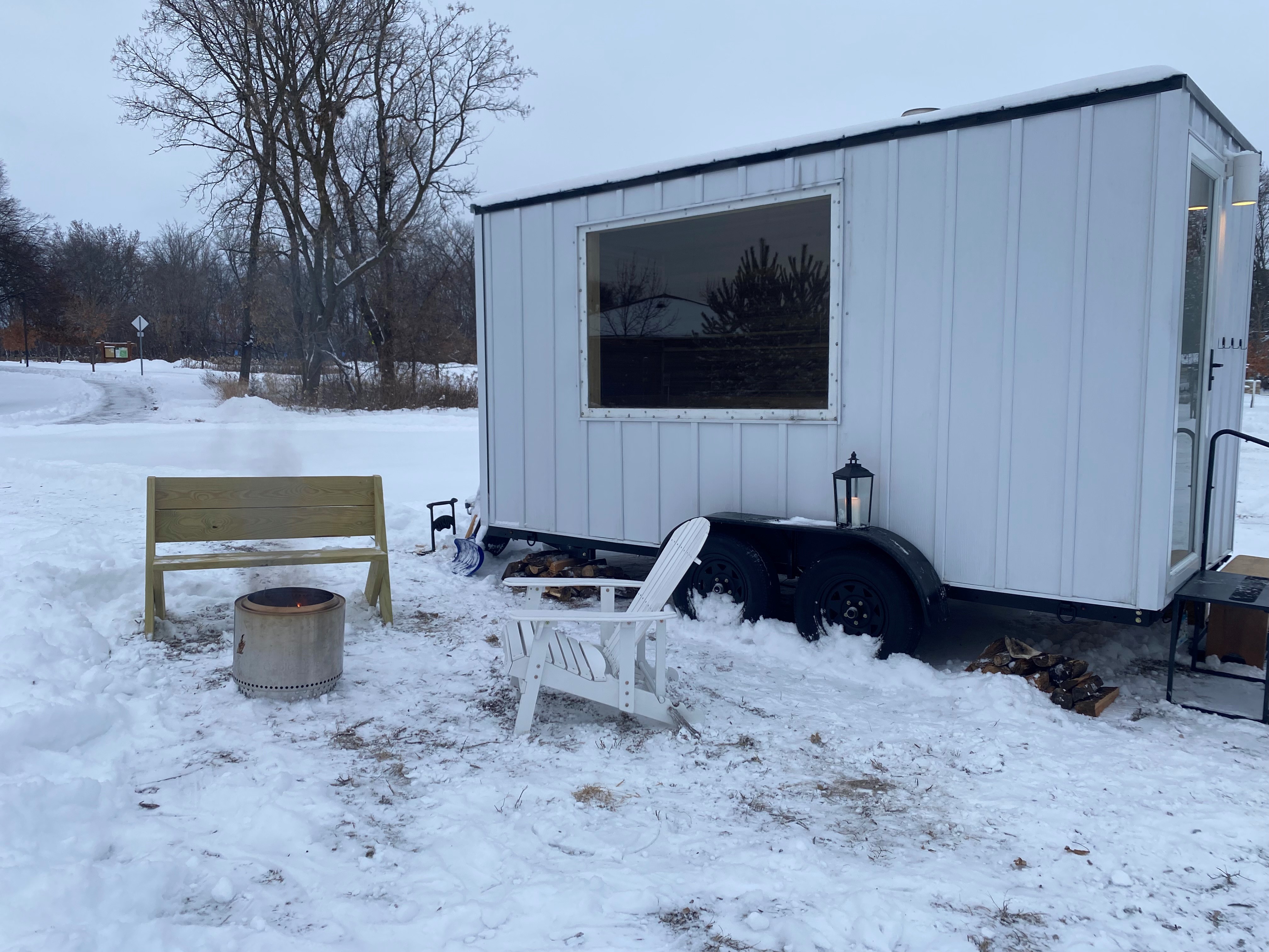 A white metal mobile sauna placed on top of snow in the winter. Alongside there are chairs and a small bonfire.