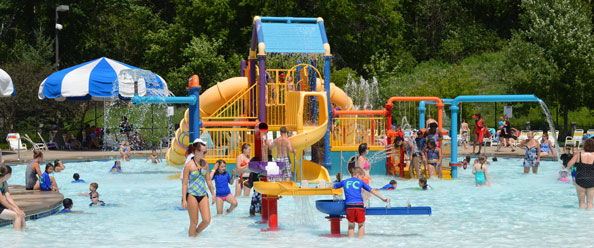 Children and adults in a wading pool with a large playground structure.